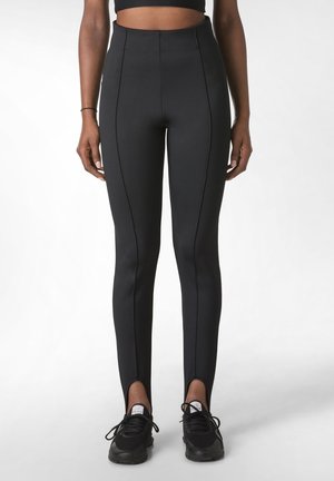 Leggings noirs taille haute avec une texture lisse et extensible, ornés de coutures contrastées et de fentes au niveau de la cheville, associés à des baskets noires.