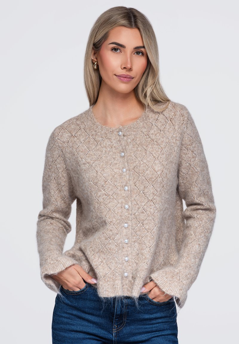 Femme aux cheveux blonds clairs portant un cardigan beige en maille avec des boutons en perles et un jean bleu, mains dans les poches, fond neutre.