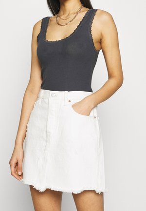 Kobieta w ciemnoszarej bezrękawniku z prążkowanym materiałem i koronkowym wykończeniem oraz białej jeansowej spódnicy z postrzępionym dołem, z jedną ręką w kieszeni spódnicy.