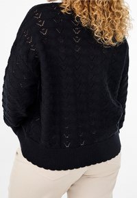 Frau mit lockigem blonden Haar, die einen schwarzen gemusterten Strickpullover und beigefarbene Hosen trägt, von hinten vor einem weißen Hintergrund gesehen.