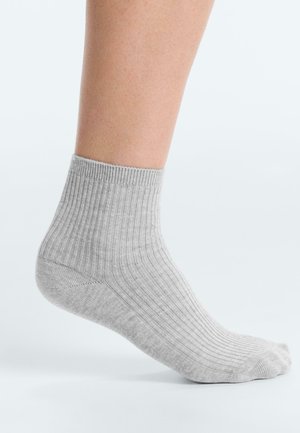 Knöchel mit einem hellgrauen, gerippten Socken, erhobener Ferse und nach unten zeigenden Zehen vor einem schlichten weißen Hintergrund.