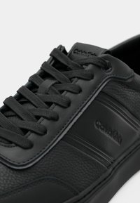 Black leather sneaker with textured finish, featuring flat laces, subtle branding, and a smooth toe cap. Stitching details enhance design.