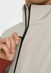 Beige Jacke mit hohem Kragen, mit schwarzem Reißverschluss-Detail. Hand greift den Reißverschluss, darunter ist ein roter Pullover sichtbar.
