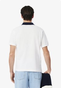 Camisa polo blanca con cuello marino, de mangas cortas y un ajuste relajado. La tela parece texturizada. Llevada con unos vaqueros de mezclilla azul claro.
