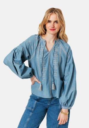 Femme aux cheveux blonds portant un chemisier bleu brodé avec des pompons et des manches longues bouffantes, associé à un jean bleu taille haute.