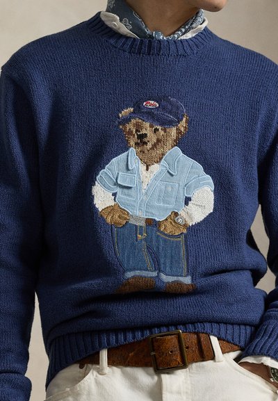 Gestrickter marineblauer Pullover mit einem bestickten Bären in Jeanshemd und Jeanshosen, der eine Mütze trägt. Gerippte Bündchen und Saum mit strukturierter Oberfläche.