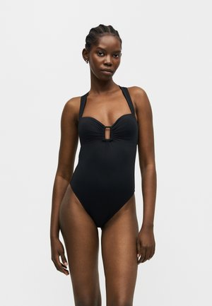 Vrouw met gevlochten haar die een zwarte eendelige badpak draagt met een uitsnede in de vorm van een sleutelgat op de borst, staand tegen een eenvoudige lichte achtergrond.