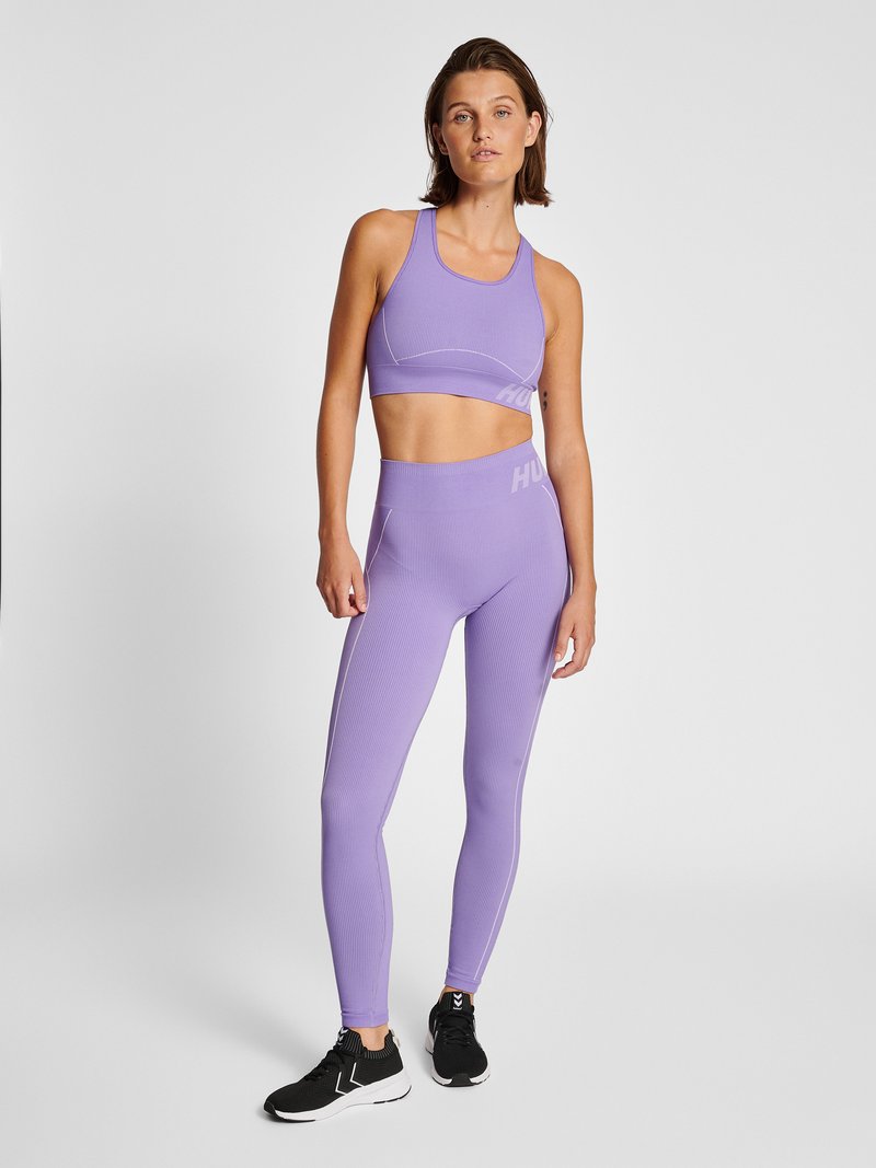 Femme debout en brassière de sport violette et leggings avec des baskets noires sur fond clair uni.
