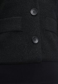 Svart texturerad jacka med två svarta knappar och sidfickor, tillverkad av ett stickat tyg med en lätt lyster och en åtsittande passform nertill.