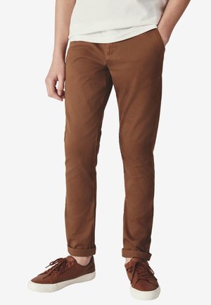 Person iført op rullede brune bukser og matchende brune sneakers med hvide såler, stående med den ene hånd i lommen.