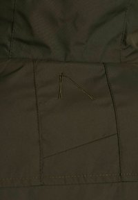 Tejido en un verde oliva oscuro con una textura suave, que presenta detalles de costura y un pequeño acento bordado en forma de triángulo en la superficie.