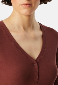 Roestkleurige, ribbelgekleurde katoenen top met een v-halslijn en drie knoopaccenten. De textuur lijkt zacht en iets rekbaar.