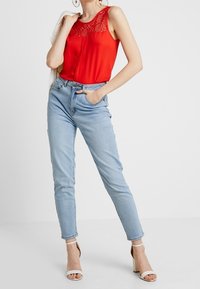 Czerwona bezrękawnik z koronkowymi detalami, zestawiony z jasnoniebieskimi obcisłymi dżinsami i białymi sandałami na słupku. Prosty design, gładka tekstura.