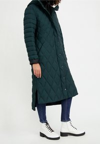 Abrigo largo acolchado en azul verdoso oscuro con capucha, cierre de botones y aberturas laterales. Combinado con botas blancas hasta el tobillo y vaqueros azules.