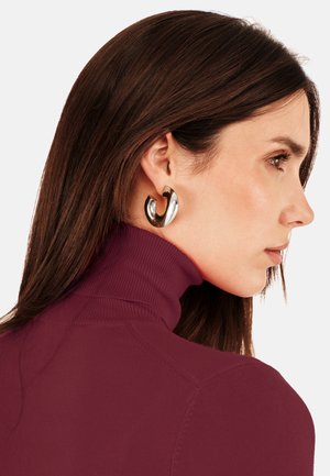 Silberne Creolen, die an einem Modell präsentiert werden, mit einer geschwungenen Form und reflektierender Oberfläche, kombiniert mit einem burgunderfarbenen Oberteil mit hohem Kragen.