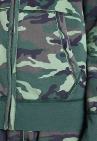 Sudadera verde con estampado de camuflaje, con cremallera, bolsillo frontal y puños acanalados. Presenta diferentes tonalidades de verde y púrpura en la tela.