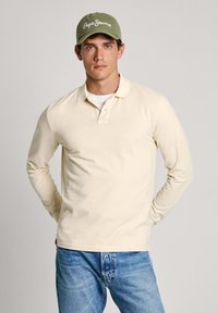 Gara piedurkne bežs polo krekls, kas izgatavots no kokvilnas, ar apkakli un diskrētu logotipu. Apvienots ar gaiši ziliem džinsiem un zaļu cepuri.