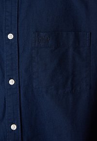 Marineblå knappskjorte laget av vevd stoff, med en brystlomme med preget "GAP"-logo og hvite knapper.