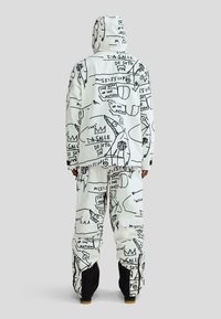 Biały zimowy komplet kurtki i spodni z czarnymi graficznymi szkicami i wzorami tekstowymi; luźny krój, z teksturowanymi mankietami i kapturem.