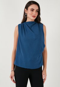 Modrý bezramínkový top s draped výstřihem a texturovanou látkou. Silueta je volná, doplněná černými těsnými kalhotami.