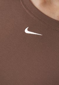 Braun gerippter Stoff mit Rundhalsausschnitt. Weißes Nike-Logo, das auf der linken Brustseite positioniert ist. Nahaufnahme zeigt die Textur des Materials.