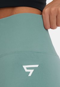 Groene sportleggingen met een geribde tailleband, een soepele stofstructuur en een prominent wit logo aan de zijkant.