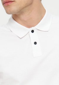 Primo piano di un uomo che indossa una polo bianca con colletto e due bottoni neri abbottonati all’altezza del collo.