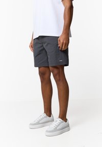 Άνδρας που φοράει σκούρα γκρι σορτς, άσπρα αθλητικά παπούτσια και λευκό πουκάμισο, στέκεται μπροστά σε απλό λευκό φόντο.