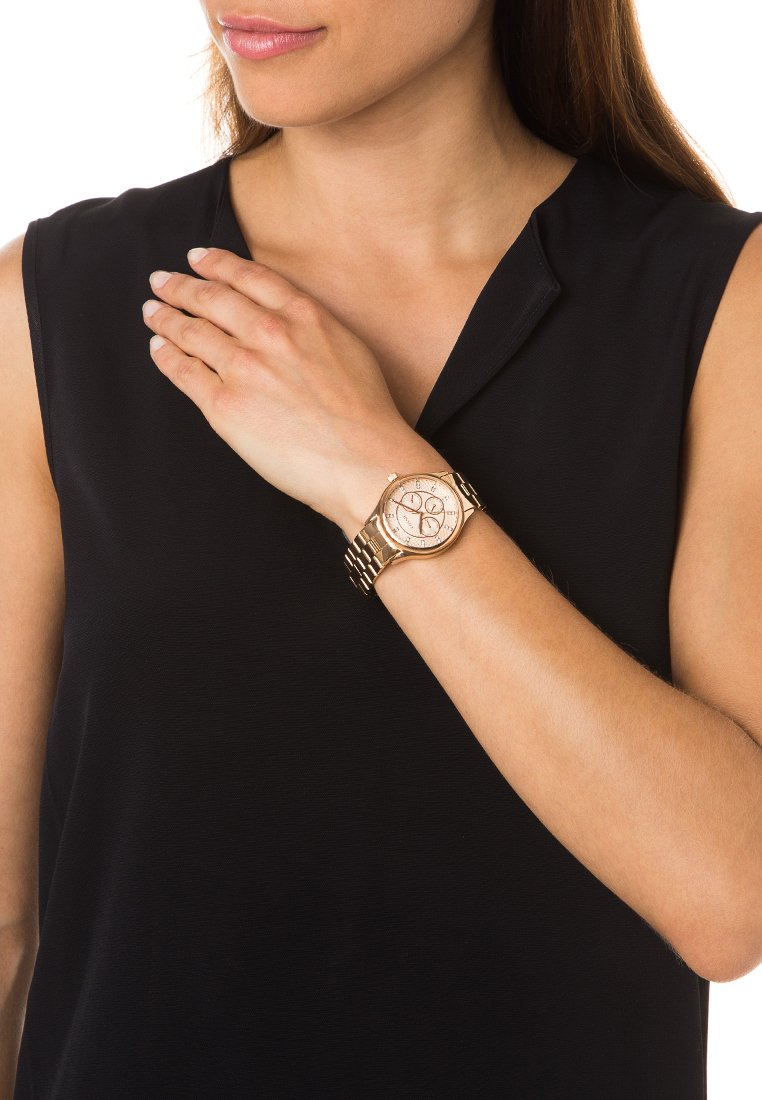 Montre en or rose avec un cadran texturé, un visage rond et un bracelet en maillons métalliques. Portée au poignet sur un haut sans manches noir, mettant en avant les détails de conception.