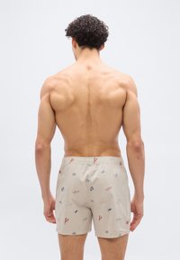 Άνδρας χωρίς πουκάμισο με σγουρά σκούρα μαλλιά γυρισμένος προς τα πίσω, φορούσε ανοιχτόχρωμα σορτς με κόκκινα και μπλε ναυτικά σχέδια.