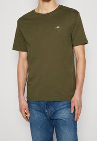 Maglietta a maniche corte verde oliva realizzata in un tessuto morbido, con un piccolo logo sul petto a sinistra e un colletto girocollo. Abbinata a jeans blu.