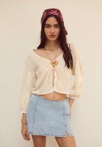 Kremast vrhnjem s V-izrezom, sprednjim vrvico in dolgimi rokavi. Svetlo modra denim mini krilo s stranskimi razporki. Naglavni zlati verižni nakit.
