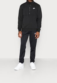 Man som bär en svart Nike zip-up sweatshirt, svarta Nike sweatpants och vita sneakers, stående mot en enkel ljus bakgrund.