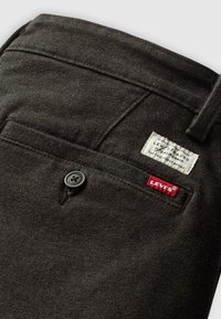Poche arrière d'un pantalon en tissu gris foncé avec un bouton noir, une étiquette de marque blanche et une étiquette logo Levi's rouge sur fond blanc.
