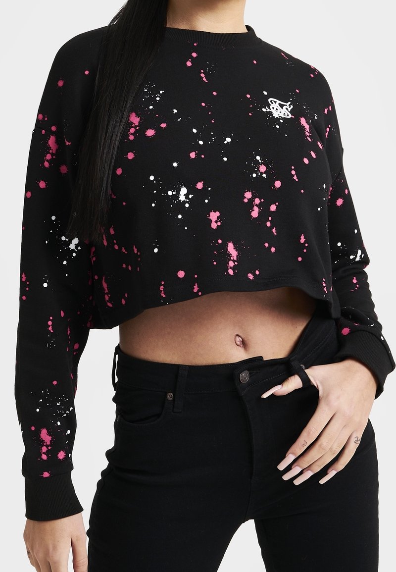 Sweat-shirt court noir avec motif éclaboussures de peinture rose et blanche, associé à un jean noir, montrant le ventre et une main ajustant la taille.