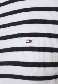 Tessuto a coste a righe bianco e navy con un piccolo logo ricamato in rosso, bianco e blu. La vista ravvicinata mette in evidenza la texture e il motivo.