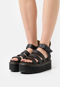 Sandales à plateforme en cuir noir avec plusieurs brides et une boucle. La semelle épaisse présente une finition texturée et des coutures jaunes contrastantes.
