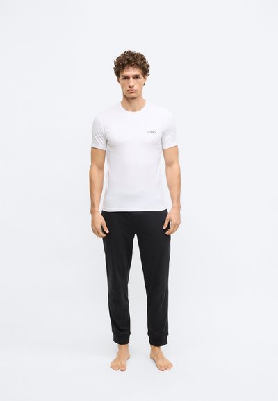 Balta īsām piedurknēm pieguloša T-krekla ar mazu logotipu uz krūtīm, kombinēts ar melnām pieskaņotām biksēm ar elastīgām aprocēm.