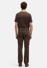 Mężczyzna o kręconych włosach, ubrany w brązowy t-shirt i pasujące brązowe jeansy, stoi tyłem na tle jednolitej białej ściany.