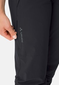 Pantaloni da esterno leggeri e scuri con una texture liscia, dotati di una tasca laterale e del logo "Vaude" in grigio. Progettati per un uso attivo.