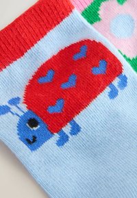 Bunte Socken mit einem roten Marienkäfer und Herzen auf einem hellblauen Hintergrund, vollendet mit einem strukturierten roten Rippbündchen und floralen Mustern.