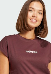 T-shirt in cotone color bordeaux con maniche arrotolate e logo "adidas" bianco centrato sul petto. Texture liscia, design casual.