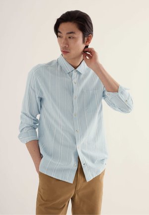 Joven con camisa de botones a rayas azul claro con las mangas remangadas, pantalones beige, de pie con una mano detrás del cuello y la otra en el bolsillo.