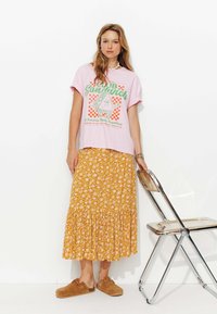 T-shirt graphique rose portant le texte "Club Sandwich", associé à une jupe maxi florale jaune et des sabots marron. Se tenant à côté d'une chaise métallique.