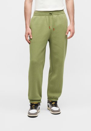 Bărbat purtând pantaloni jogger verde măsliniu cu talie cu șnur și manșete elastice, asortați cu adidași albi și gri cu șireturi negre.