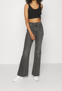 Kvinna i svart kort topp, högmidjade urvuxna svarta wide-leg jeans och vita sneakers som står mot en enfärgad bakgrund.