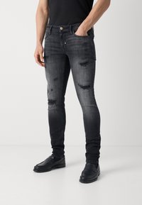 Jeans de mezclilla negra con parches desgastados y áreas descoloridas, diseño ajustado, estilo de cinco bolsillos y cierre de botón. Combinados con zapatos oscuros.