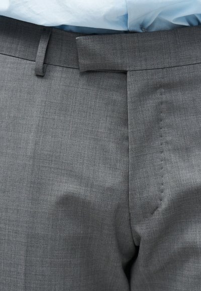 Γκρι παντελόνι με κούμπωμα και θηλιές για ζώνη, φορεμένο με ένα γαλάζιο πουκάμισο που έχει μπει μέσα στο παντελόνι.