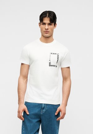 Mężczyzna w białej koszulce z krótkim rękawem z czarnym logo Karl Lagerfeld Jeans na piersi, ubrany w niebieskie dżinsy, stojący na jednolitym tle.