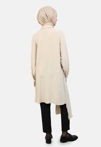 Beige long tunic featuring a textured fabric, long sleeves, and a high-low hem. Paired with black pants and black shoes.